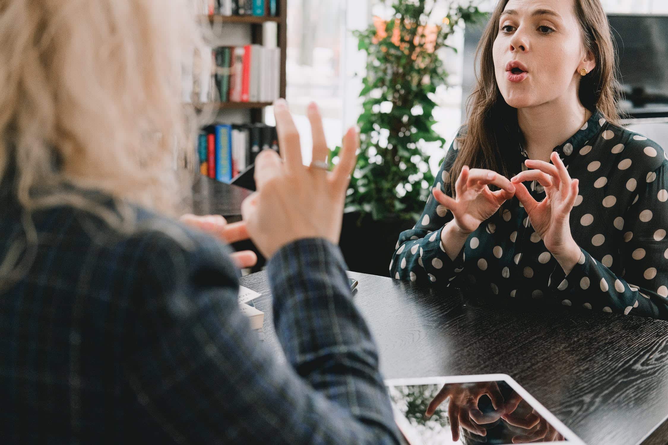 A photo of two women sat at a table, using British Sign Language to speak with each other.