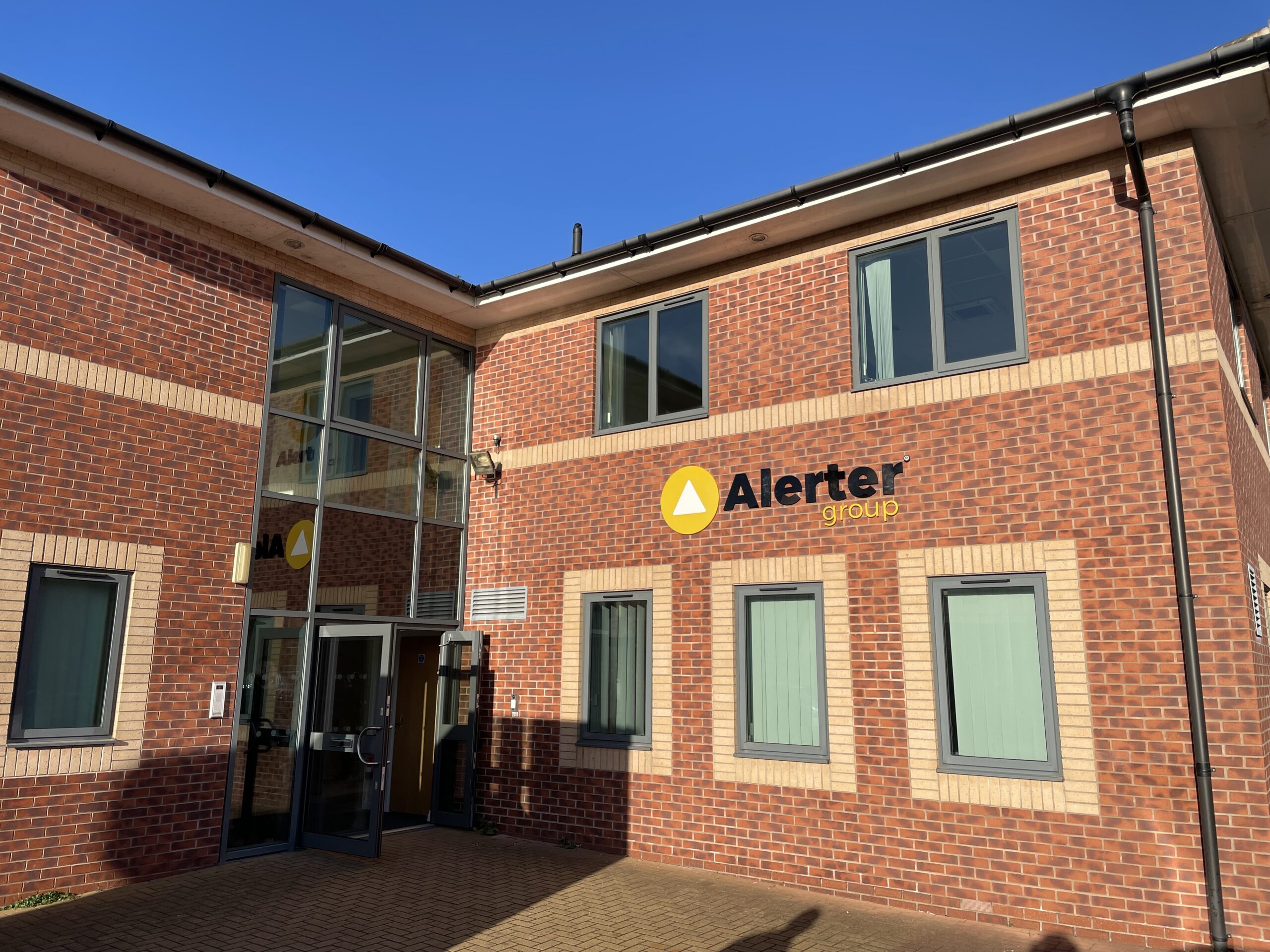 A photo of the outside of Alerter Group's head office. The building is constructed from red brick, and the company logo is on the front of the building. The front doors are open and it's a sunny day with clear blue skies.